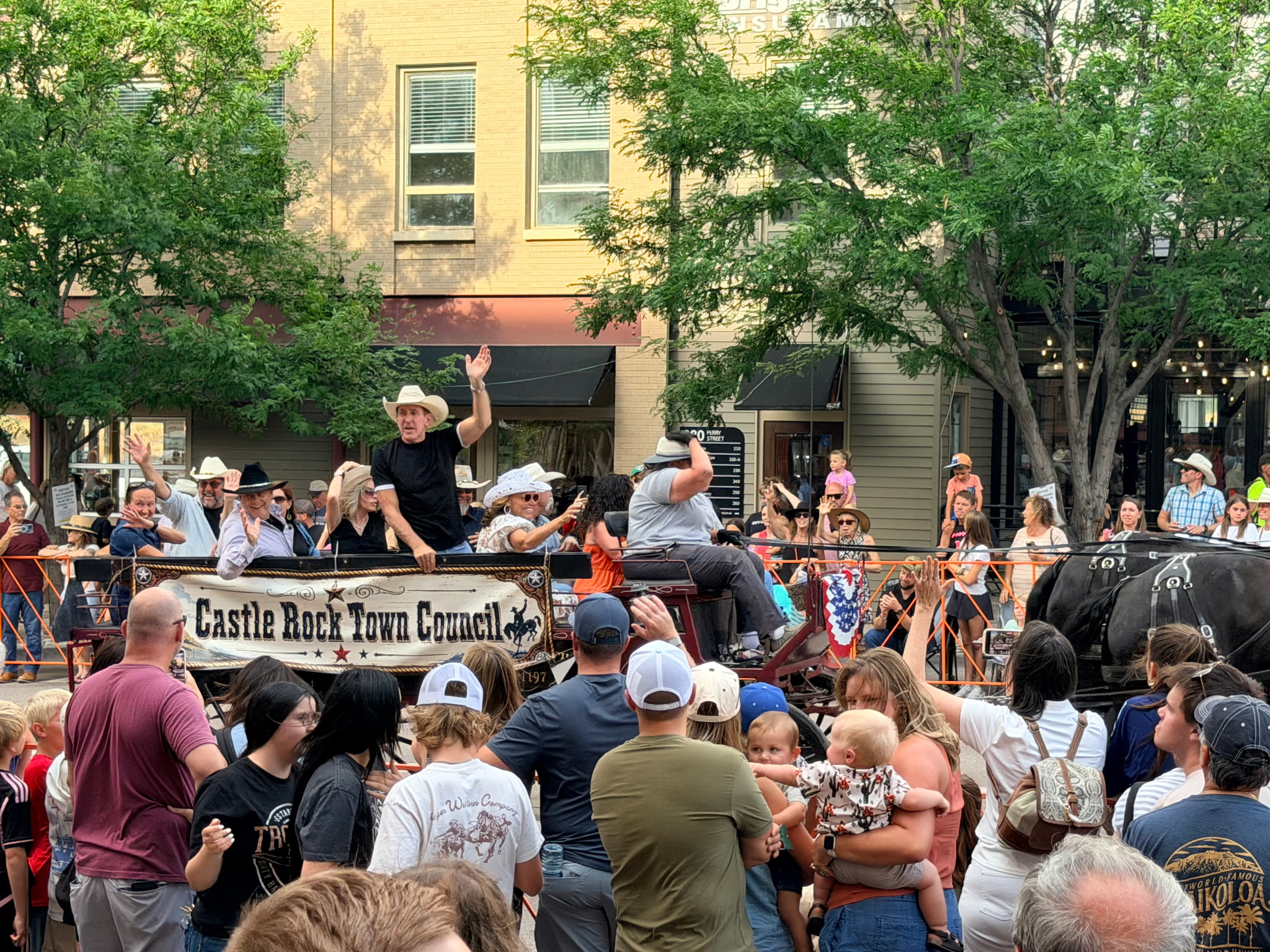 Tim Dietz standing and waving from the Castle Rock Town Council wagon as the Western Heritage Cattle Drive rolls down Perry Street past crowds in downtown Castle Rock
