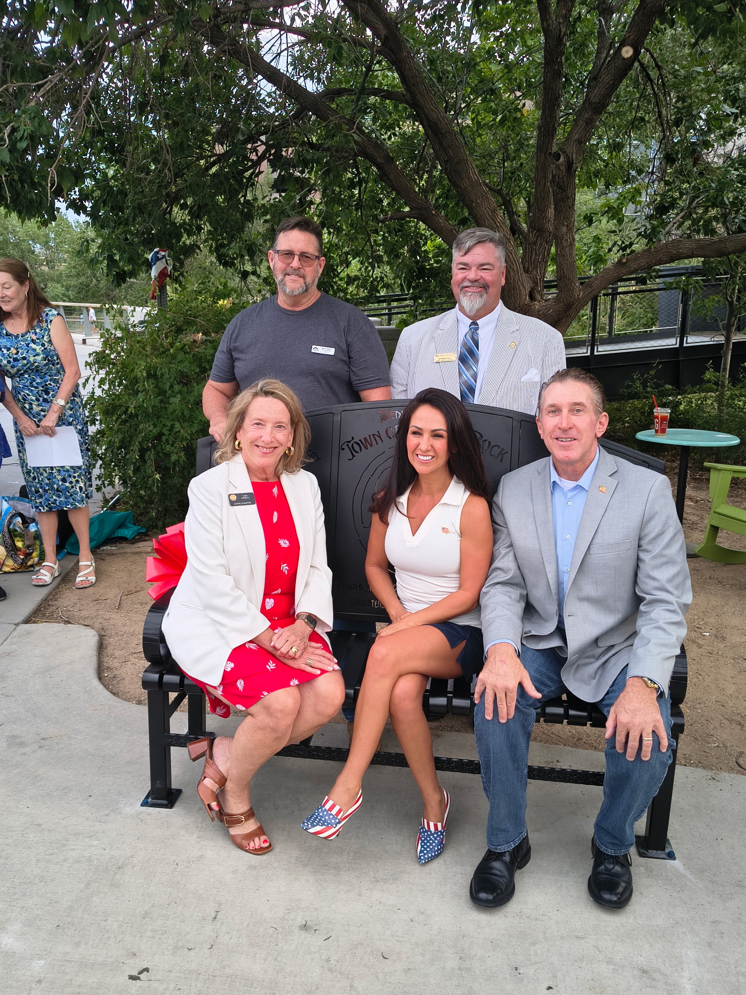 Tim Dietz seated on the newly dedicated veterans bench at Festival Park beside U.S. Representative Lauren Boebert and State Senator Lisa Frizell, with local officials gathered around and a red dedication ribbon on the bench