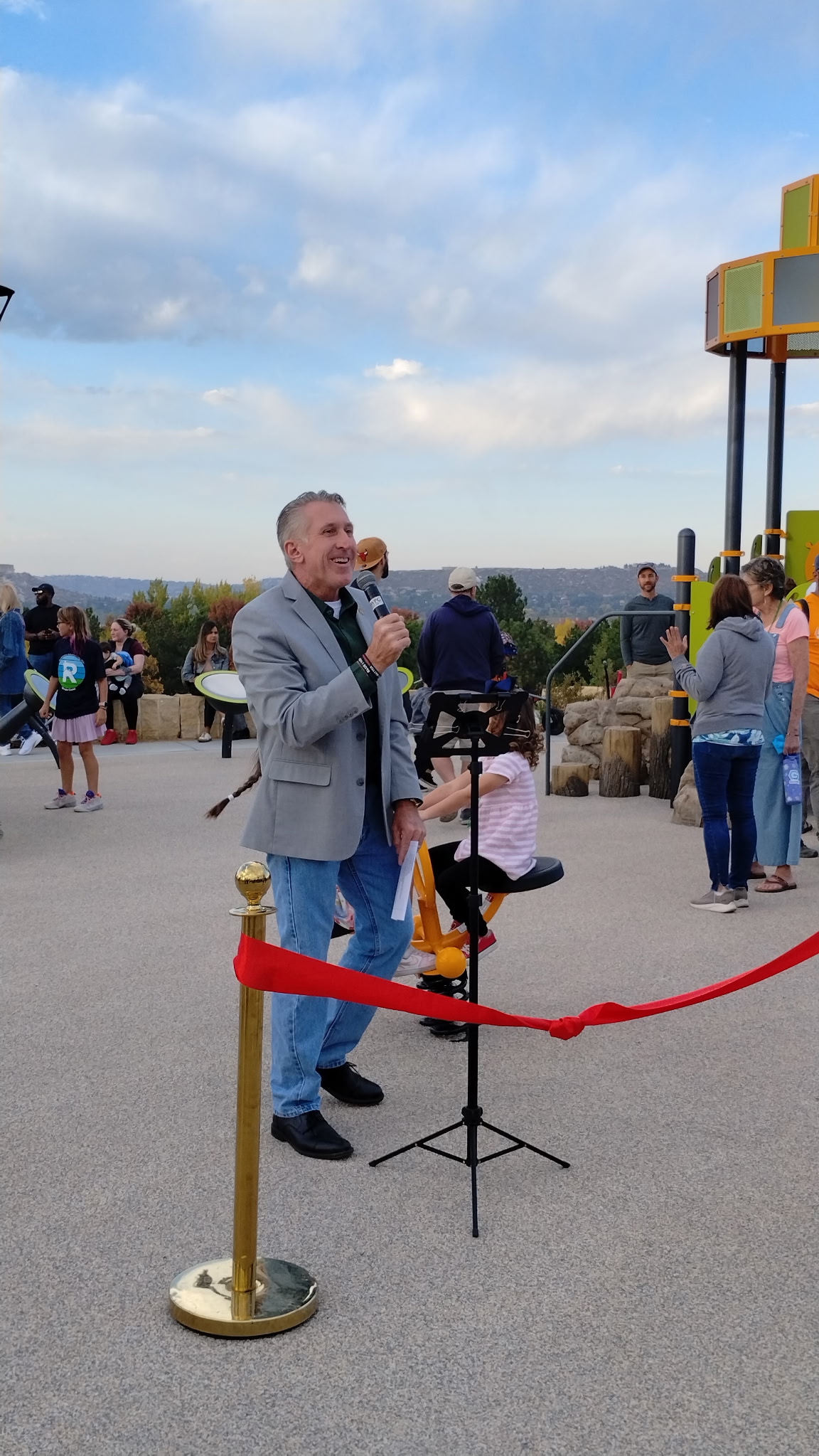 Tim Dietz speaking into a microphone at the Emerald Park grand opening in Castle Rock, behind the red dedication ribbon with families and kids on the new playground behind him