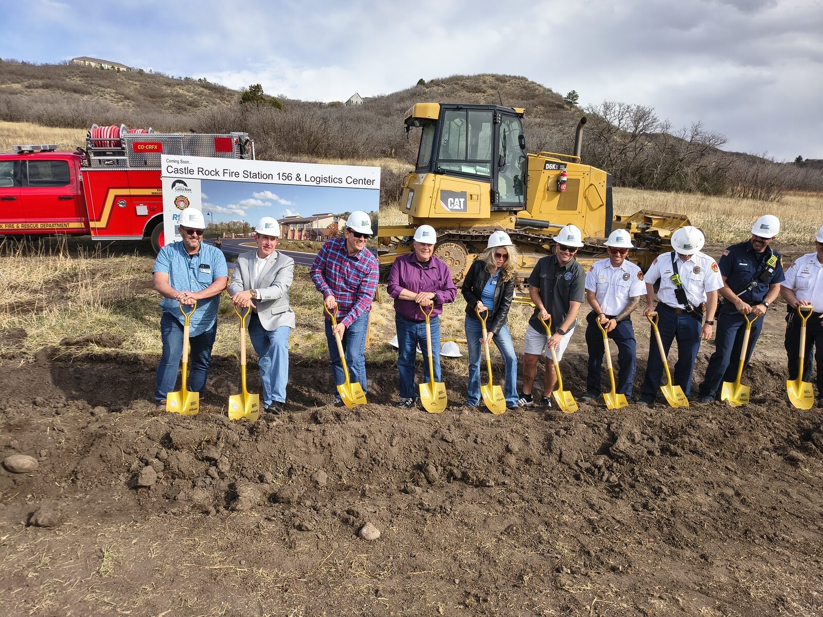 Town officials and Castle Rock Fire and Rescue leadership in white hard hats turning dirt with gold shovels at the Fire Station 156 groundbreaking, with a fire truck and bulldozer behind them
