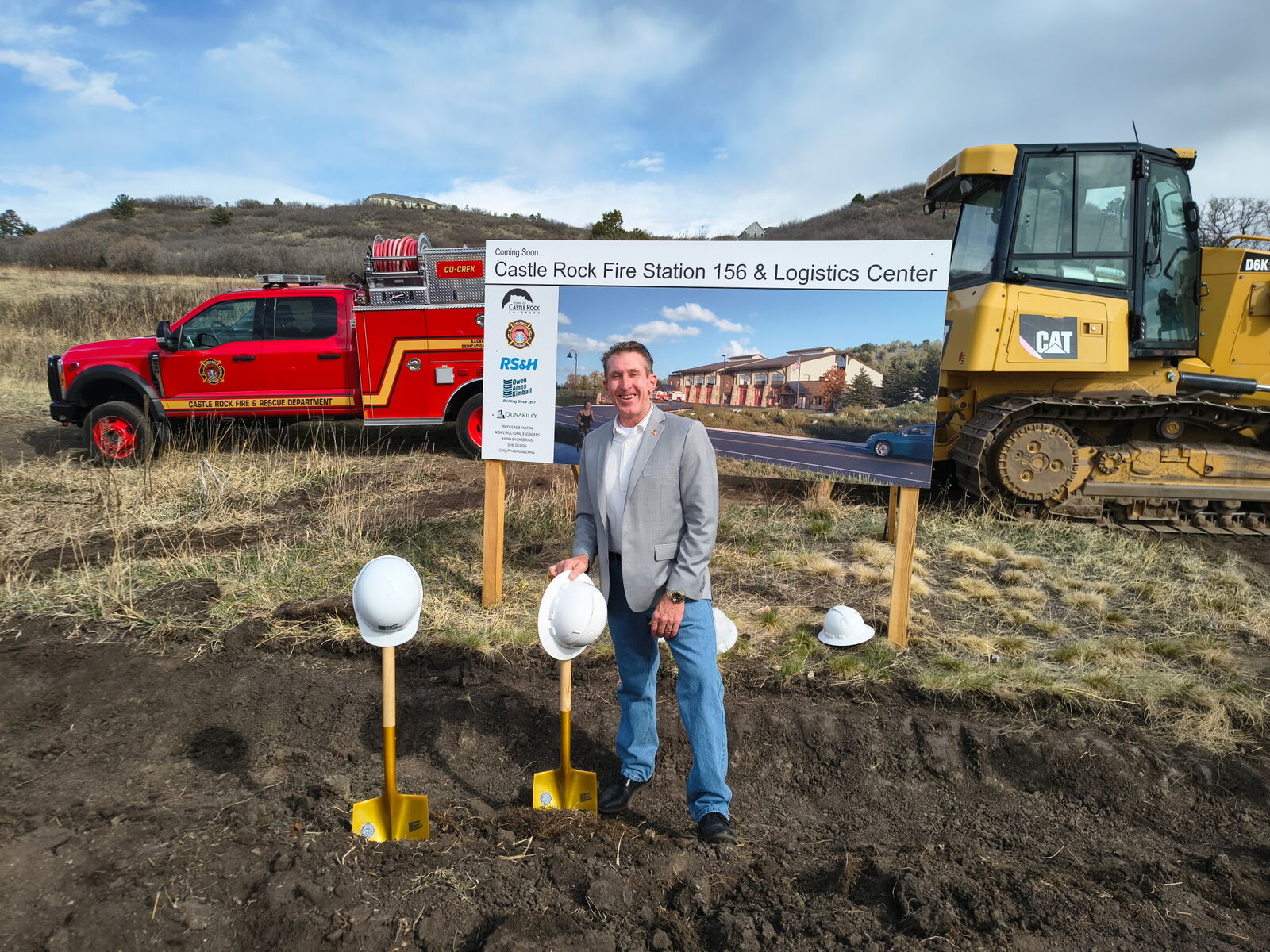 Tim Dietz in a hard-hat-and-shovel groundbreaking setting at the Castle Rock Fire Station 156 & Logistics Center site sign, with a Castle Rock Fire & Rescue brush truck and bulldozer behind him