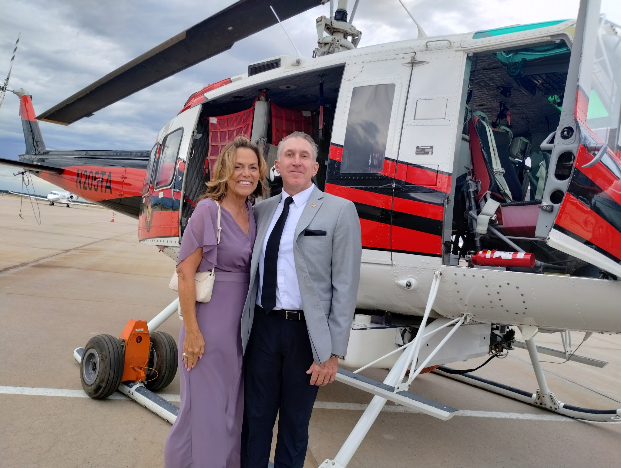 Tim Dietz in a gray suit and his wife JoAnna in a lavender gown in front of a red-and-white firefighting helicopter on the tarmac at the Douglas County Heroes Gala