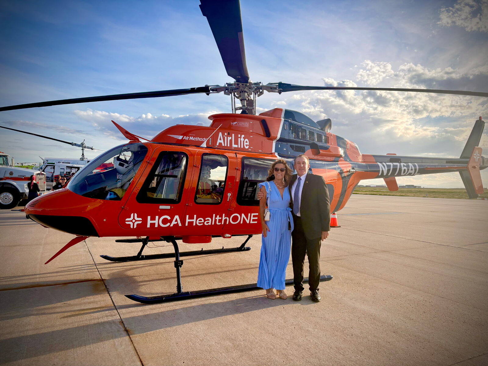 Tim and JoAnna Dietz on the tarmac beside a red HCA HealthONE AirLife medical helicopter under a big Colorado sky at Centennial Airport