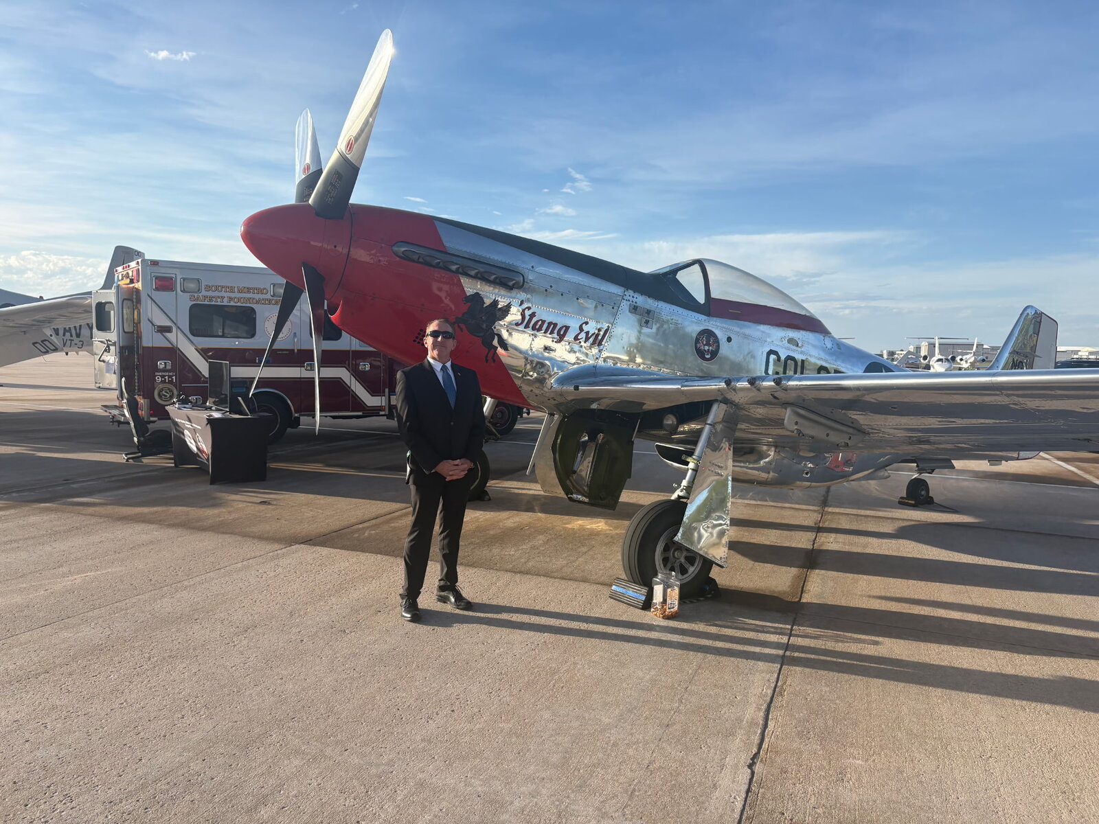 Tim Dietz in a dark suit standing in front of a polished red-and-silver P-51 Mustang named Stang Evil, with a South Metro Safety Foundation ambulance behind, on the tarmac at Signature Aviation