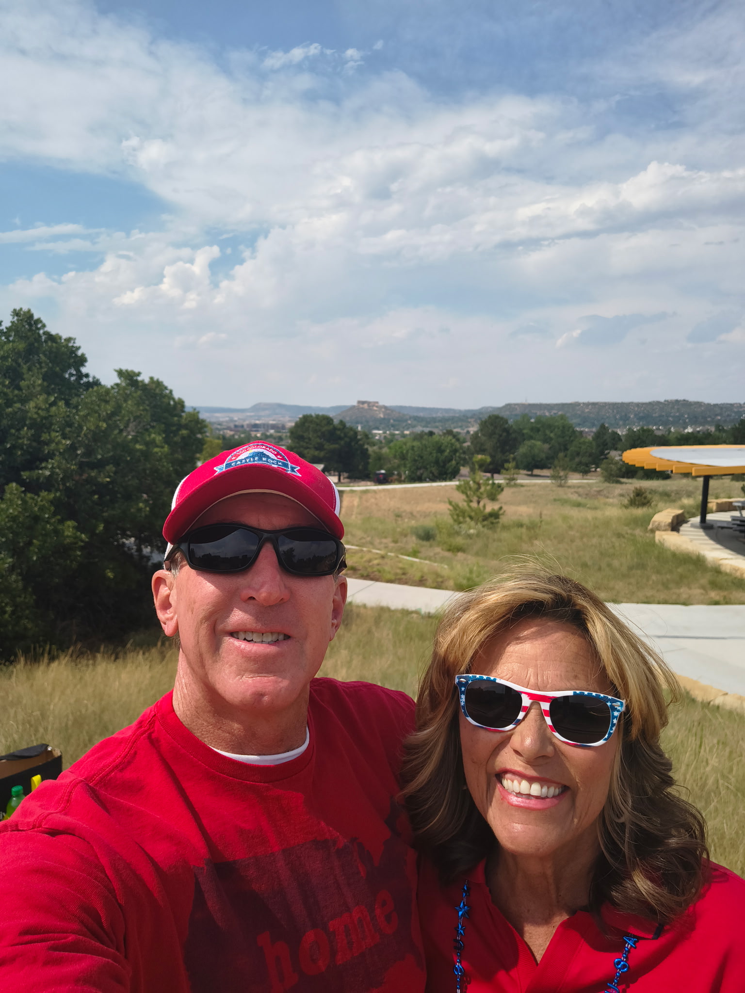 Tim Dietz in a red Castle Rock cap with his wife JoAnna in stars-and-stripes sunglasses, celebrating the Fourth of July in Castle Rock with The Rock on the horizon behind them