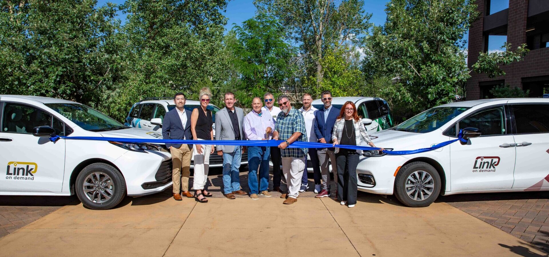 Tim Dietz with Douglas County Commissioners, Castle Rock officials, CDOT representatives, and Via Transportation at the Link On Demand ribbon cutting, holding a blue ribbon in front of the new branded rideshare vans