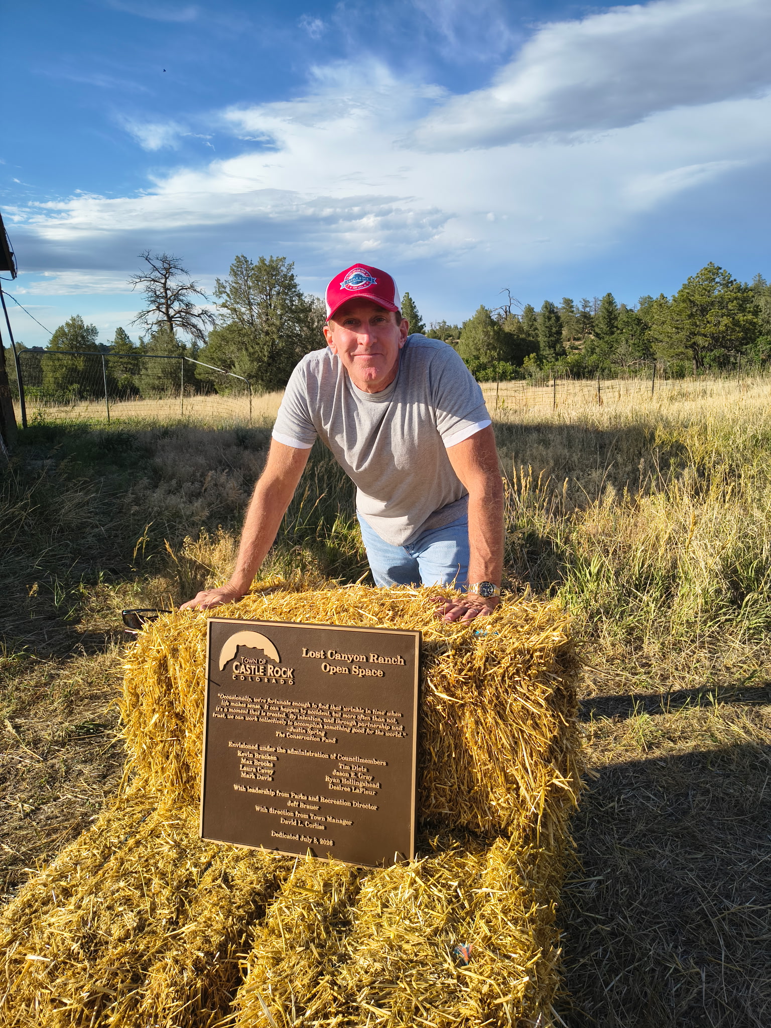 Tim Dietz at the Lost Canyon Ranch Open Space dedication, leaning over the bronze Town of Castle Rock plaque set on hay bales, with grasslands and pines behind him