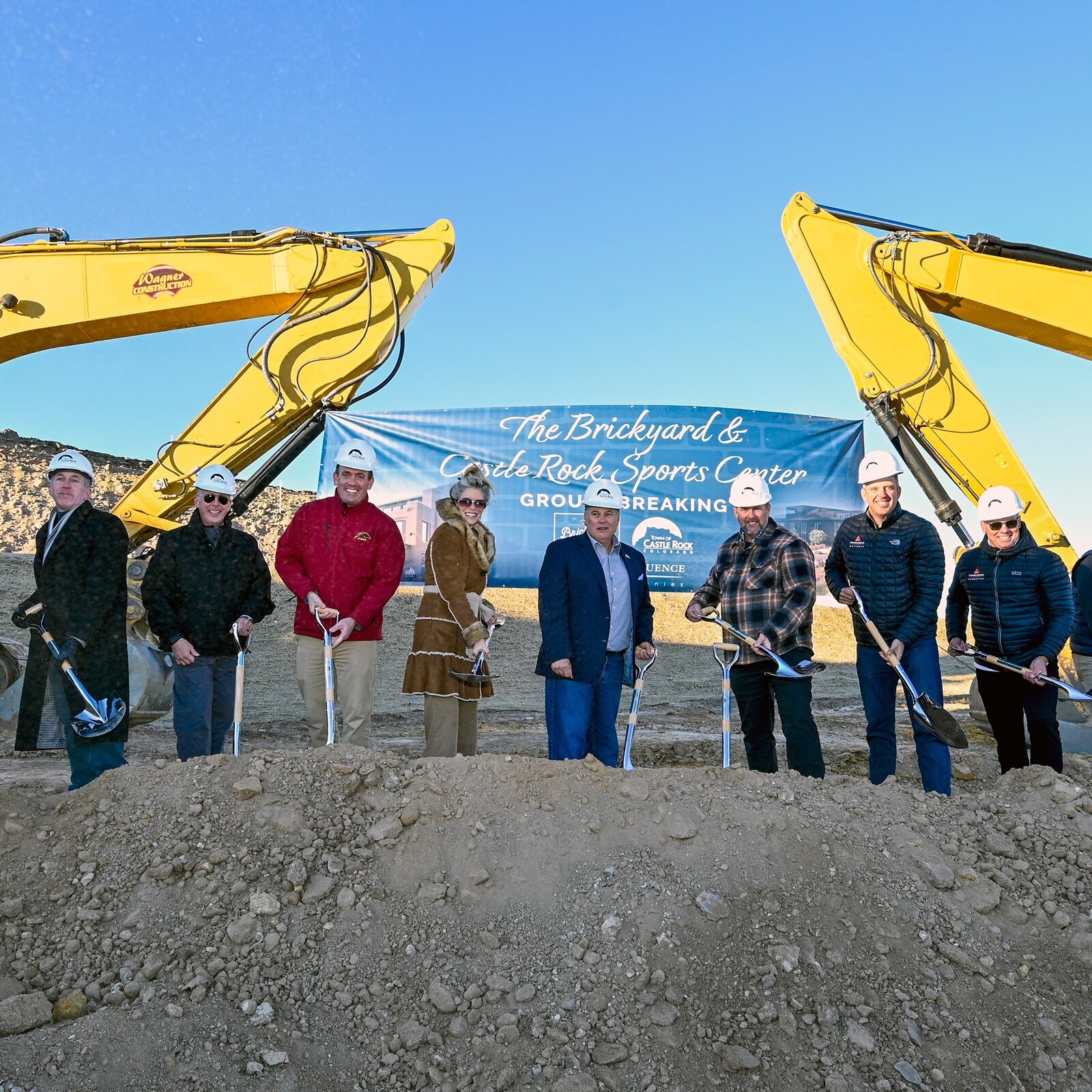 Tim Dietz and Laura Cavey with Town leaders and project partners in hard hats, turning dirt with silver shovels between two excavators at the Brickyard and Castle Rock Sports Center groundbreaking
