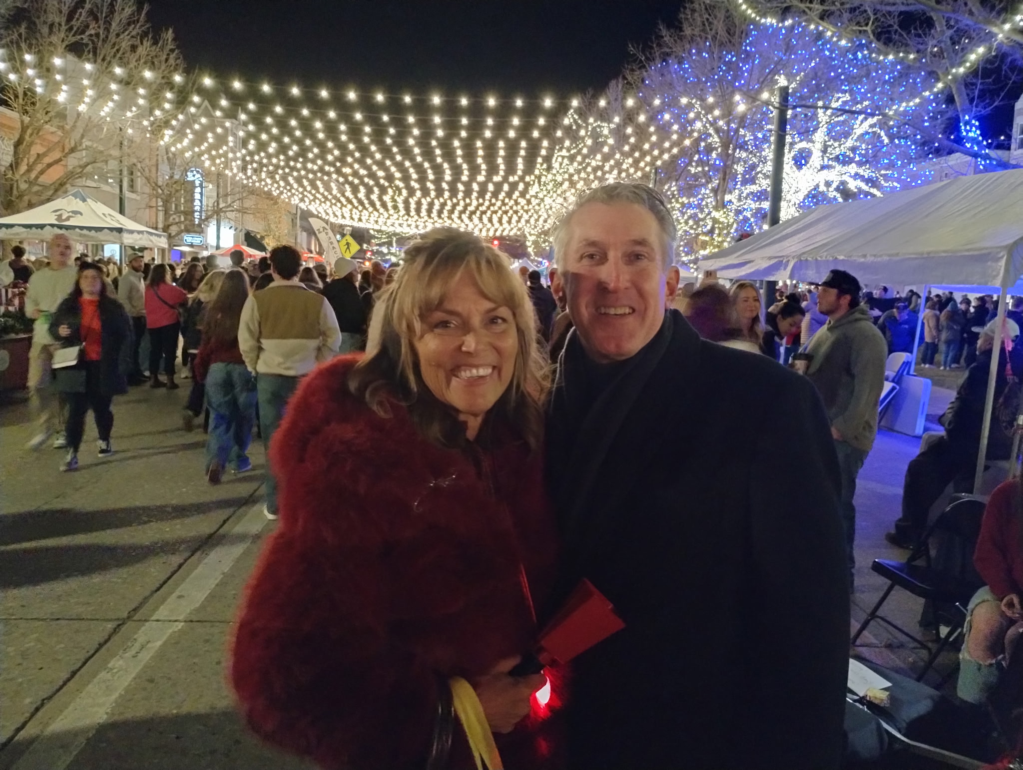 Tim Dietz and his wife JoAnna at the Castle Rock Starlighting, smiling under a canopy of string lights in downtown Castle Rock with lit trees and crowds of neighbors behind them